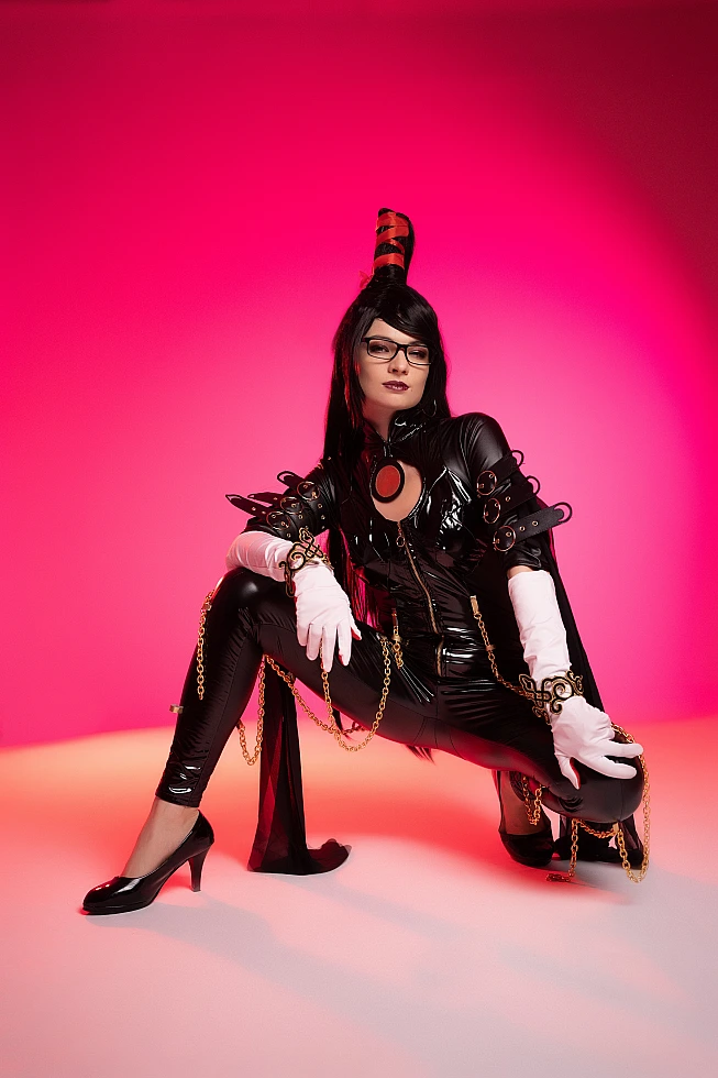 Bayonetta cosplay asymmetrical grounded knee pose by Neka-Chi, glossy black bodysuit with gold chains, saturated pink studio