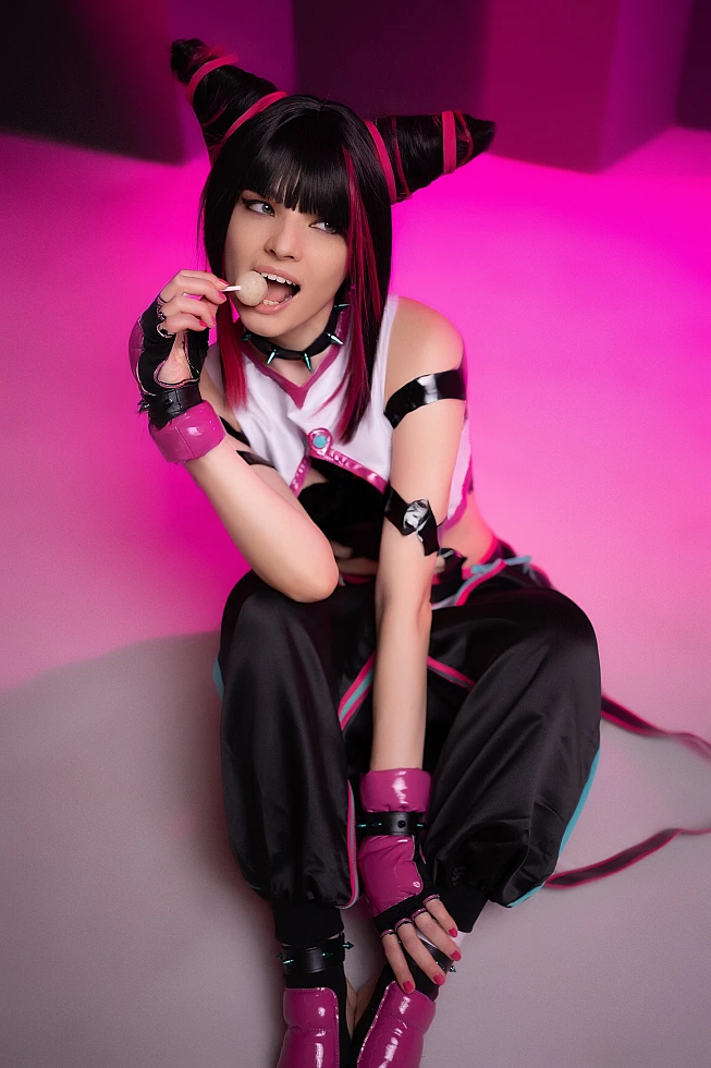 Juri Han cosplayer sitting on studio floor with candy near lips, magenta neon lighting, glossy strap costume, horn buns hairstyle
