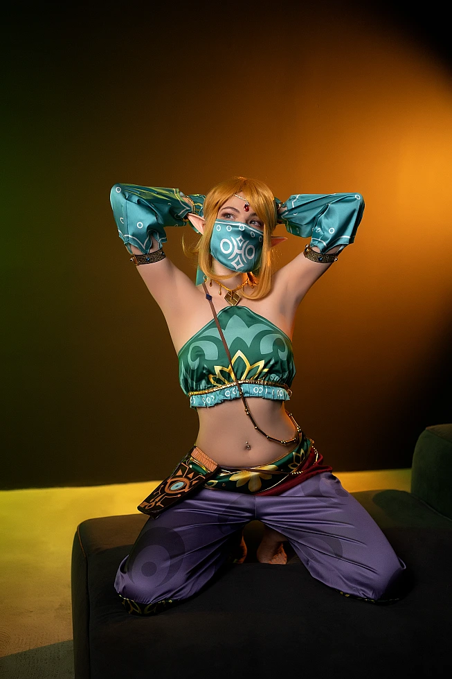 Gerudo Link cosplay kneeling pose with veil mask, emerald outfit and amber lighting