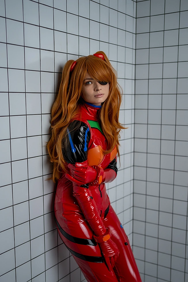 Introspective Asuka Langley cosplay leaning against tiled corner wall, muted studio lighting on red plugsuit, long hair and compact restrained pose