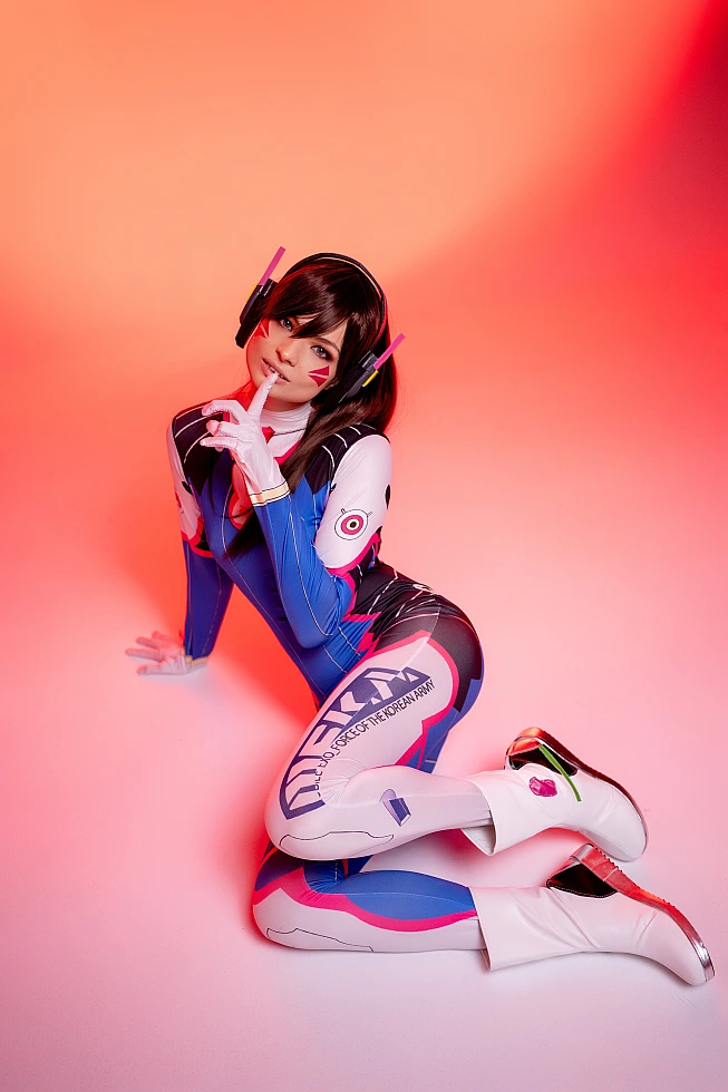 D.Va Overwatch cosplay kneeling pose finger to lips gesture blue bodysuit warm gradient studio portrait