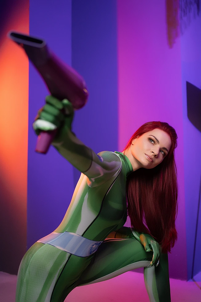 Dramatic crouching Sam Totally Spies cosplay in green catsuit with raised gadget under magenta and violet neon studio lighting low angle shot
