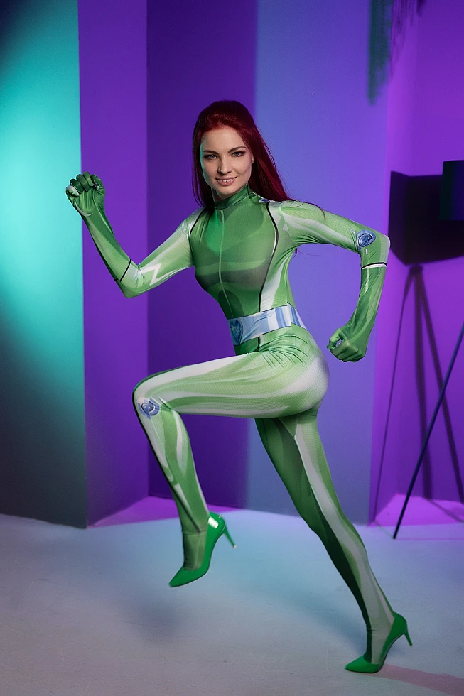 Powerful stance Sam Totally Spies cosplay with raised knee in green spy catsuit with red hair against modern geometric color panel studio background