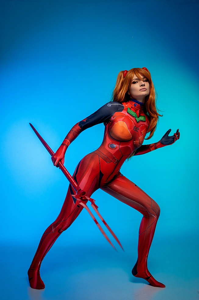 Asuka Langley cosplay lunging forward in red plugsuit with weapon against blue studio background