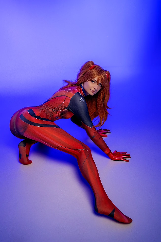 Asuka Langley cosplay crouching low in red plugsuit looking over shoulder on blue background