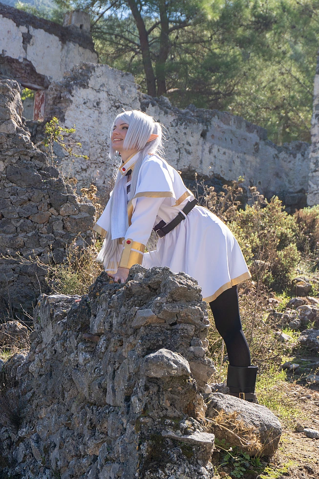 Frieren cosplay leaning low stone wall silver hair elf ears white gold mage outfit ruined architecture natural daylight gentle expression