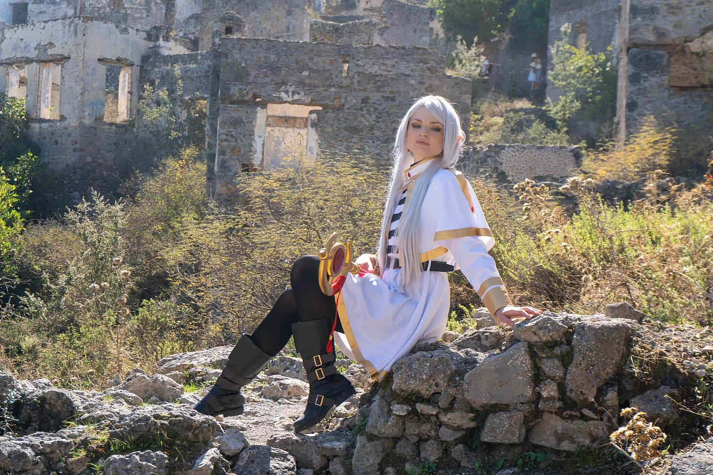 Frieren cosplay seated layered stone ruins holding staff silver hair elf ears white gold mage dress overgrown walls sunlight
