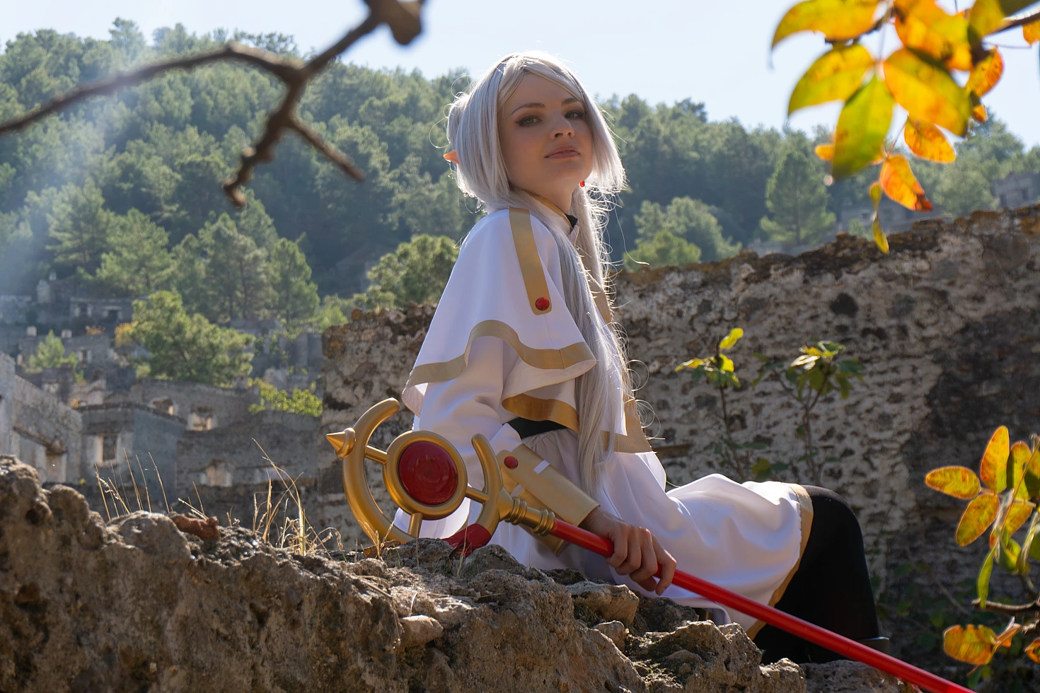 Frieren cosplay low angle seated portrait silver hair elf ears white gold dress crimson staff stone ruins forest backdrop