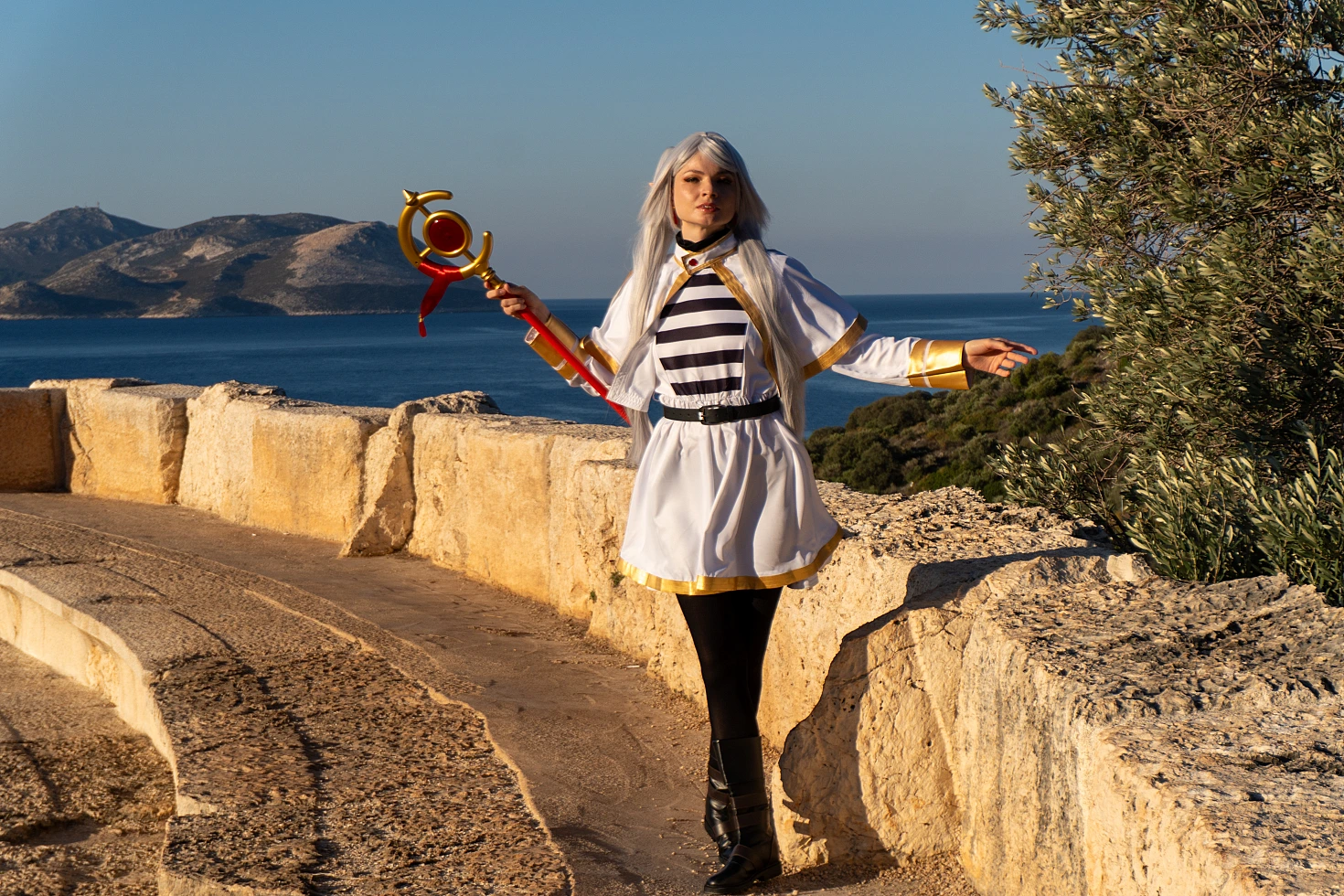 Frieren cosplay standing stone wall near sea silver hair elf ears white gold mage dress staff coastal horizon sunlight