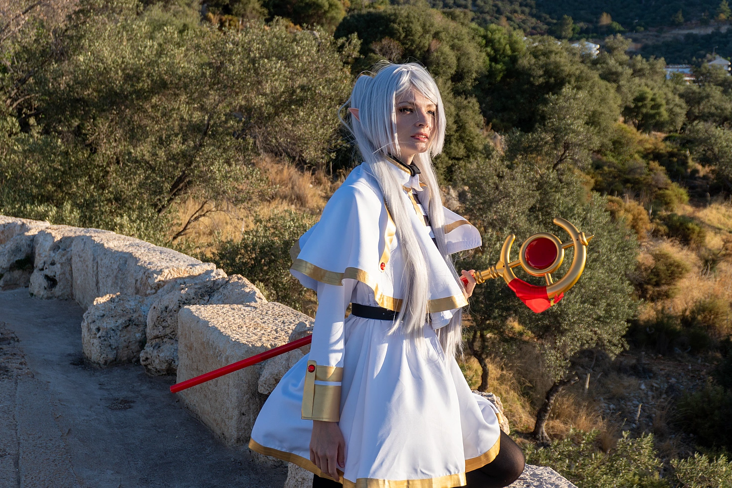 Frieren cosplay walking side profile beside stone wall silver hair elf ears white gold dress staff trailing forest hillside