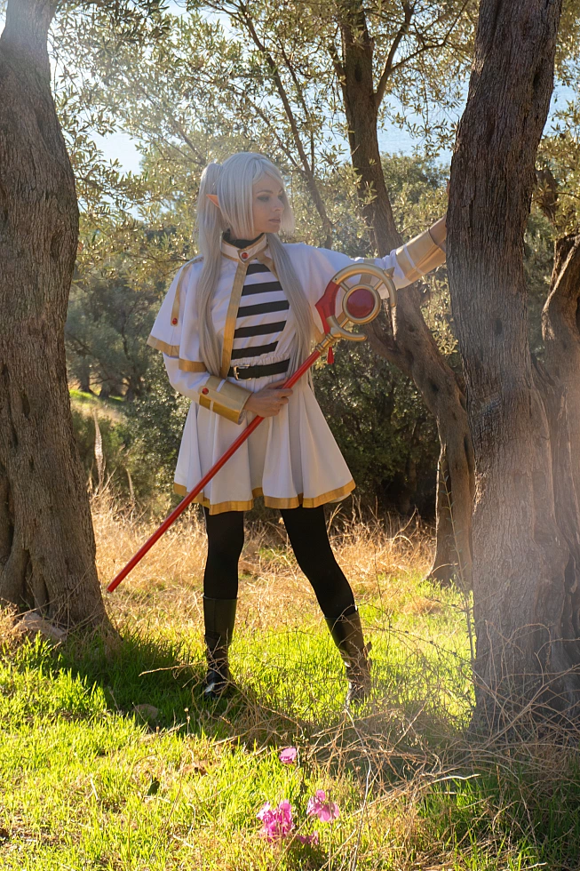 Frieren cosplay standing between two trees staff held vertically silver hair elf ears white gold mage dress forest clearing daylight