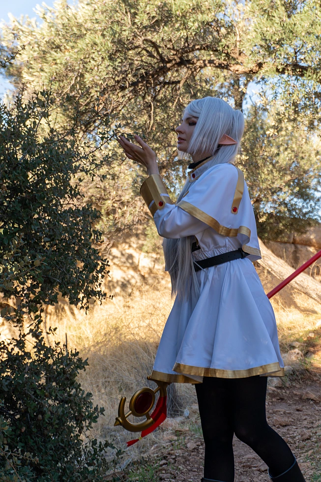 Frieren cosplay profile pose hands raised toward leaves silver hair elf ears white gold mage dress forest light staff behind