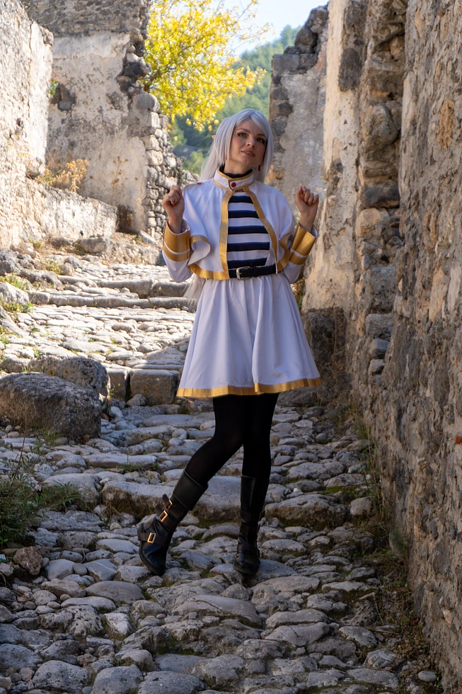 Frieren cosplay full-body pose narrow stone alley silver hair elf ears white gold mage dress cobblestones daylight