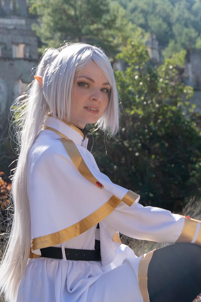 Frieren cosplay close portrait soft gaze silver hair elf ears white gold mage outfit blurred ruins and greenery
