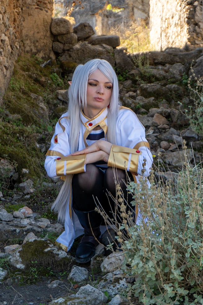Frieren cosplay crouched stone ruins downward gaze silver hair elf ears white gold mage outfit dry grass soft daylight