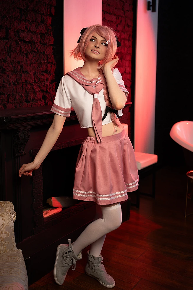 Astolfo trap cosplay standing in pink school uniform with white thigh-high socks under warm red studio lighting