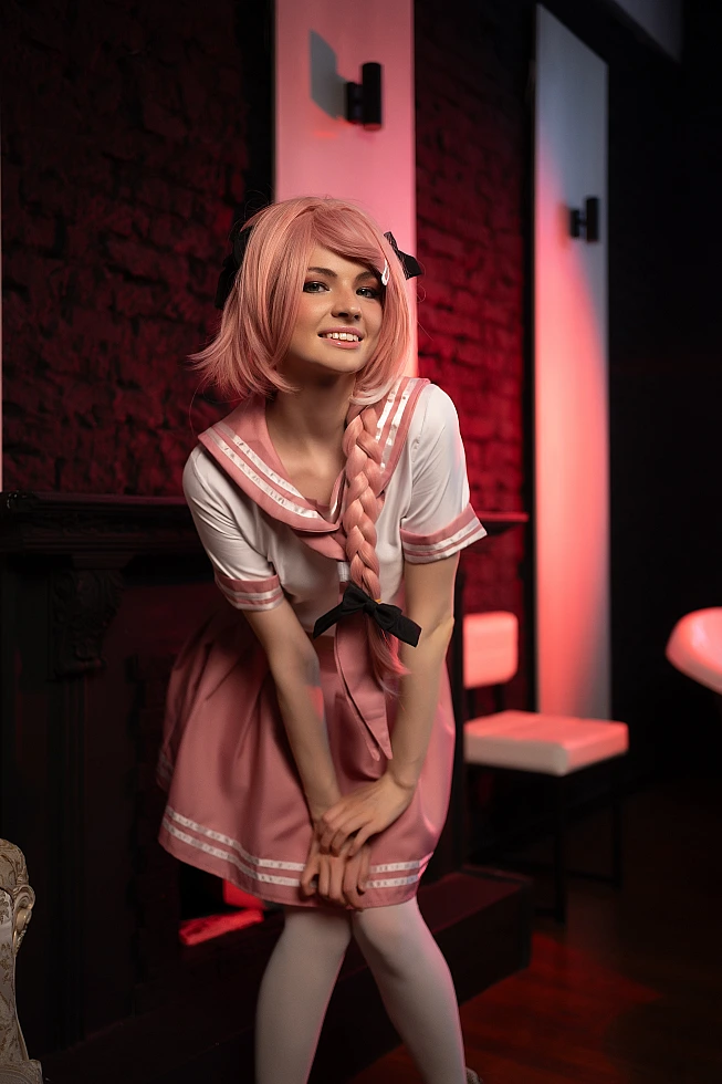 Astolfo femboy cosplay leaning forward with smile in pink school uniform and braided hair under warm red lighting