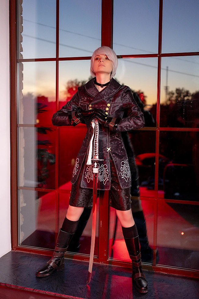 Full-body 9S cosplay standing by window at sunset with sword, black YoRHa coat and combat boots.