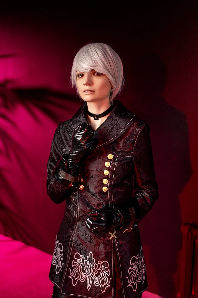 Portrait of 9S cosplay with silver wig and black YoRHa coat lit by red neon against dark background.