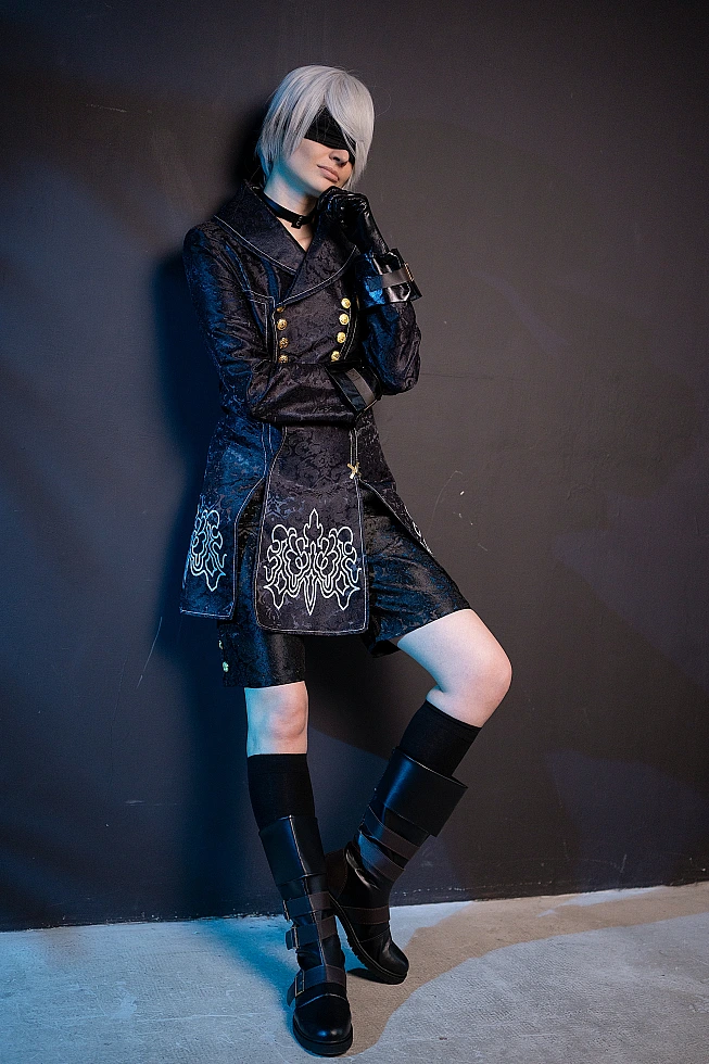 Full-body 9S cosplay from NieR:Automata in black YoRHa coat with blindfold, standing in blue rim light studio.