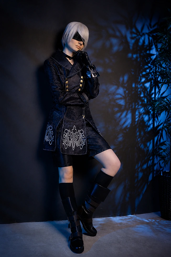 Full-body 9S cosplay from NieR:Automata in black YoRHa coat with blindfold, standing in blue rim light studio.