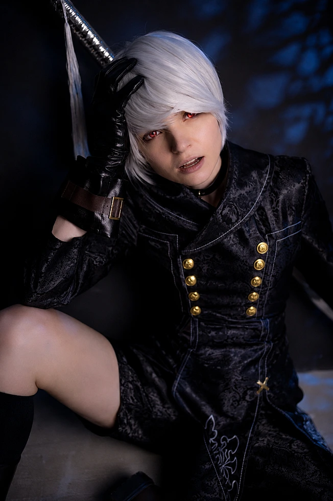 Close-up 9S cosplay portrait with silver hair, black YoRHa coat and katana hilt on dark background.