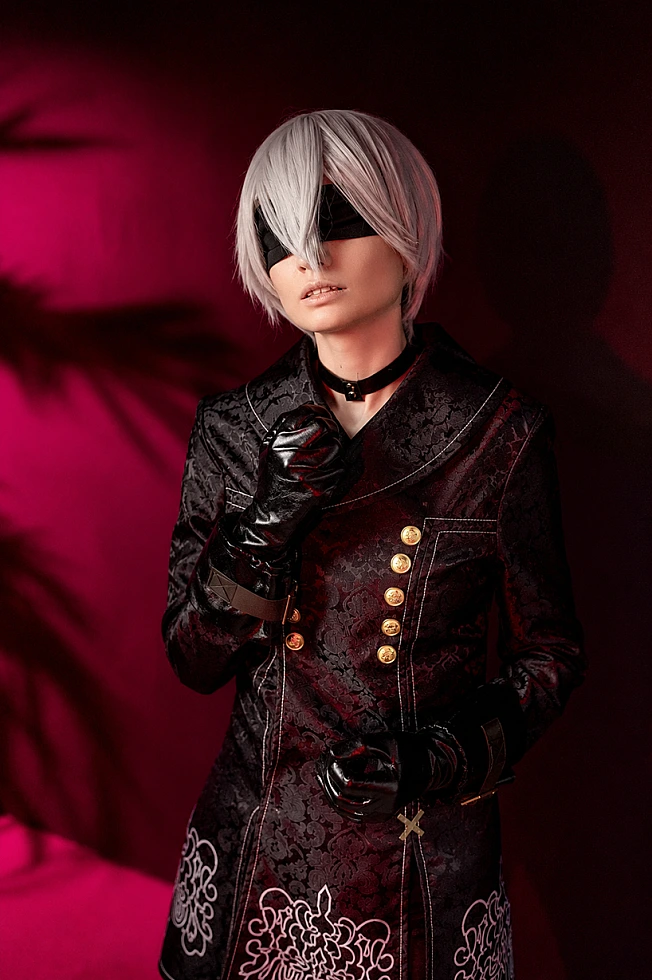Portrait of 9S cosplay with silver wig and black YoRHa coat lit by red neon against dark background.