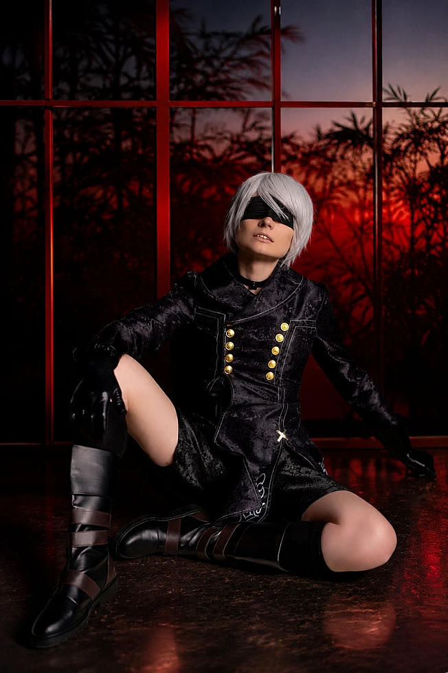Seated 9S NieR:Automata cosplay in black YoRHa coat and white wig, leaning by dark wall with sword.