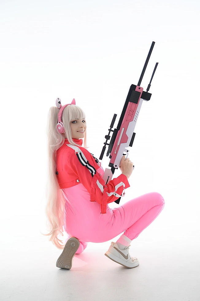 Alice cosplay from Goddess of Victory Nikke in pink tactical bodysuit holding a sniper rifle while posing in a white studio background