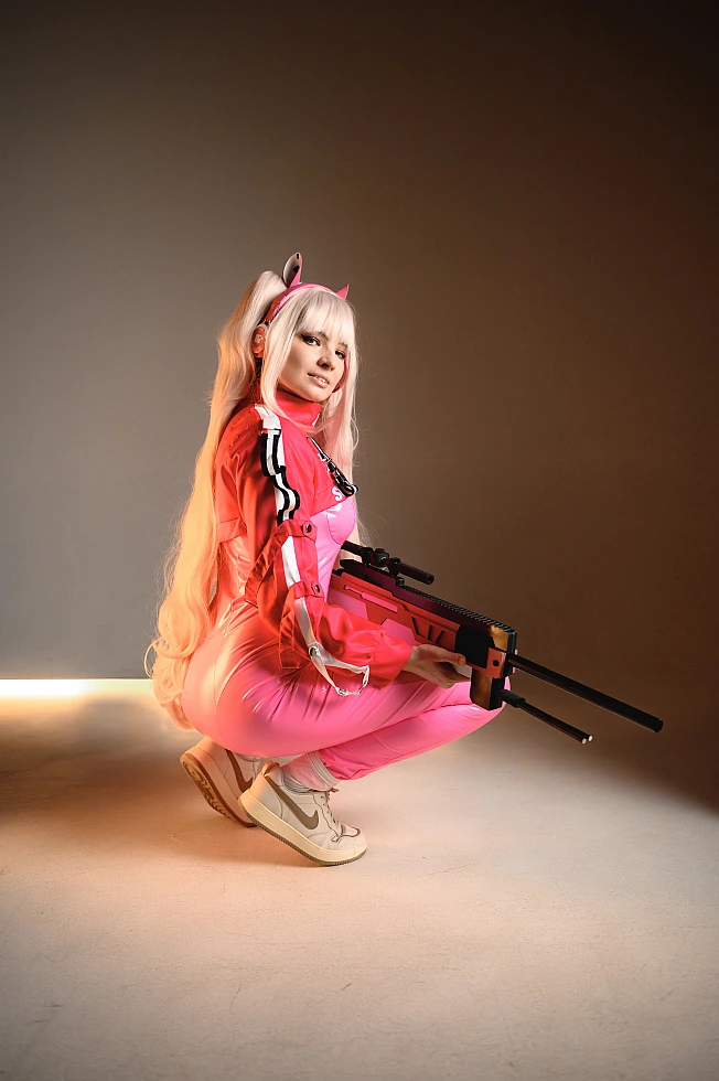 Alice cosplay from Goddess of Victory Nikke crouching with a sniper rifle under warm cinematic studio lighting