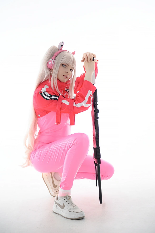 Alice cosplay from Goddess of Victory Nikke crouching beside a sniper rifle in a bright white studio