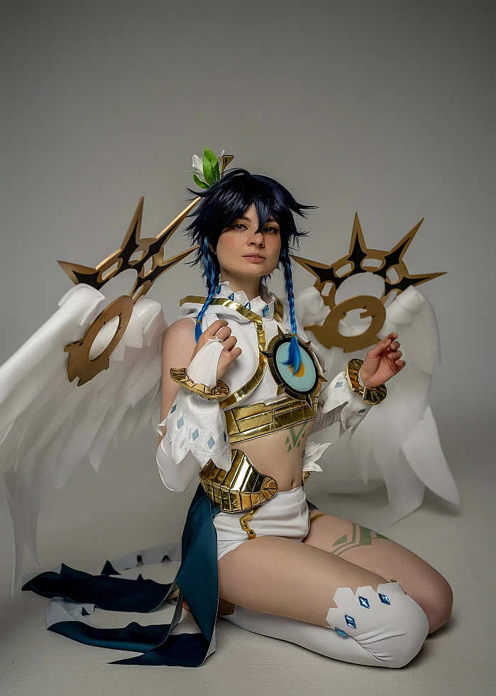 Venti cosplay from Genshin Impact kneels on a studio floor in front of large white feathered wings.