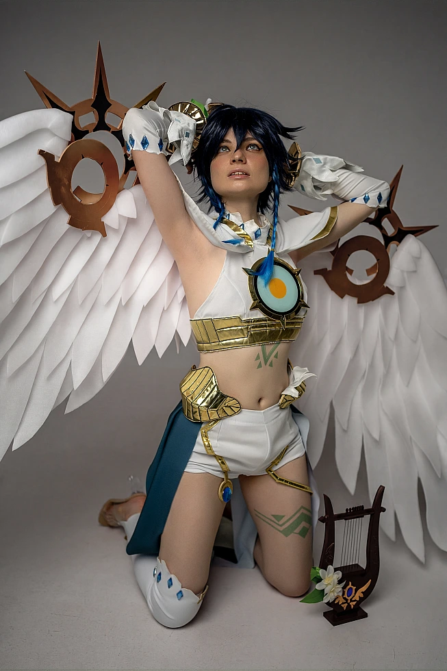 Venti cosplay from Genshin Impact kneels upright in a gray studio, white angel wings spread wide behind him.
