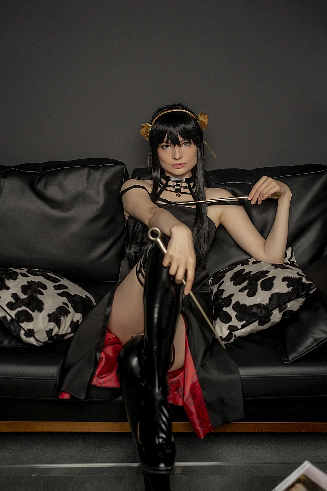 Yor Forger from Spy x Family sits on a black leather sofa, one knee raised toward the camera and a stiletto across her chest.