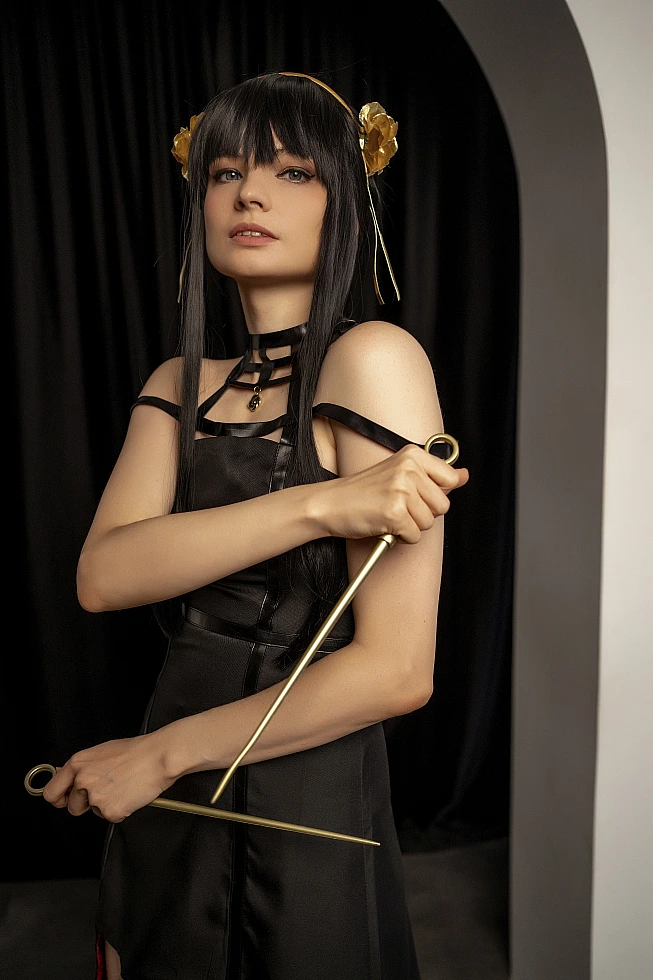 Yor Forger from Spy x Family in a half-length portrait by a black curtain, holding two gold stilettos across her body.