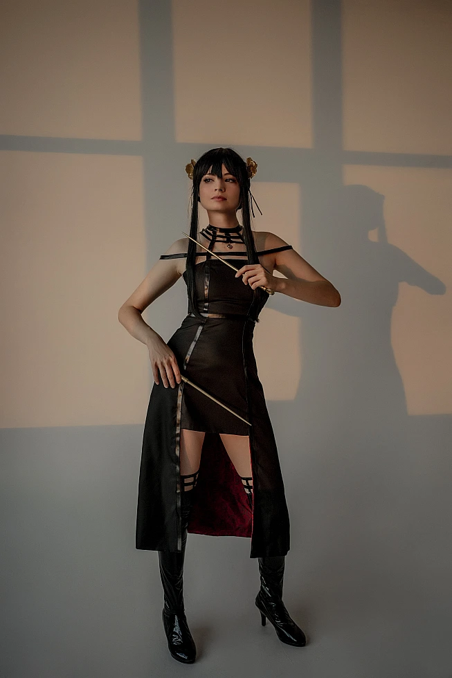 Yor Forger from Spy x Family stands full length against a warm wall crossed by window light, her shadow beside her.