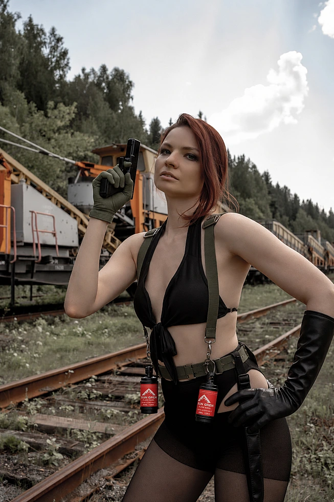 Quiet cosplay standing on rusted railway tracks with a handgun raised beside her face and orange track machines behind